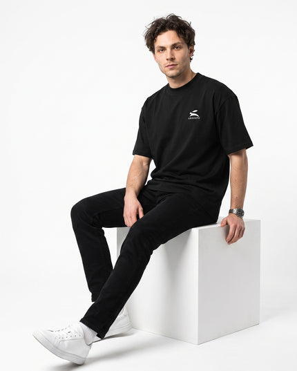Editorial streetwear studio pose of model Jax seated on minimalist white cube wearing GROZAVU Black T-Shirt in seamless white cyclorama