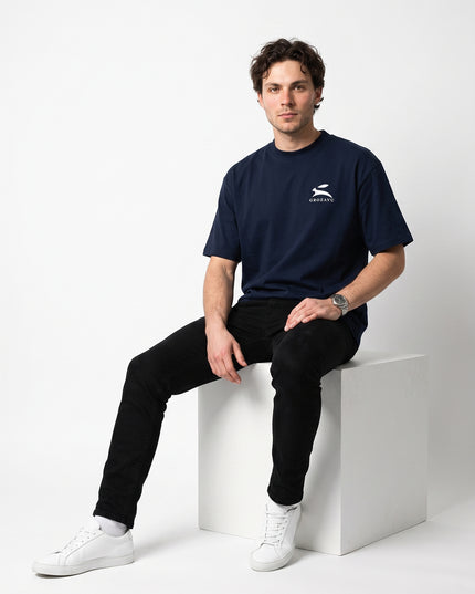 Editorial streetwear studio pose of model Jax seated on minimalist white cube wearing GROZAVU Dark Navy T-Shirt in seamless white cyclorama