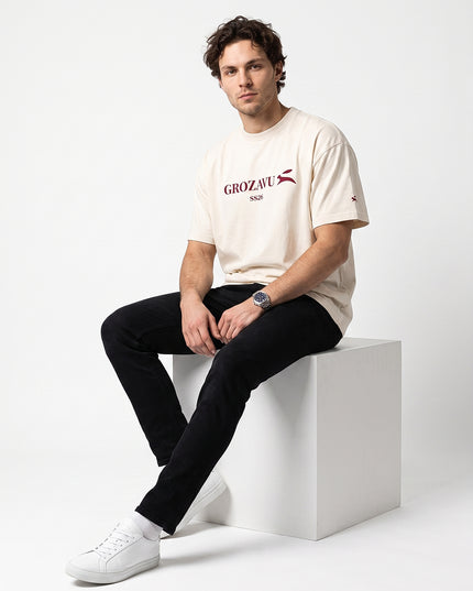 Editorial streetwear studio pose of model Jax seated on minimalist white cube wearing GROZAVU SS26 Off-White T-Shirt in seamless white cyclorama