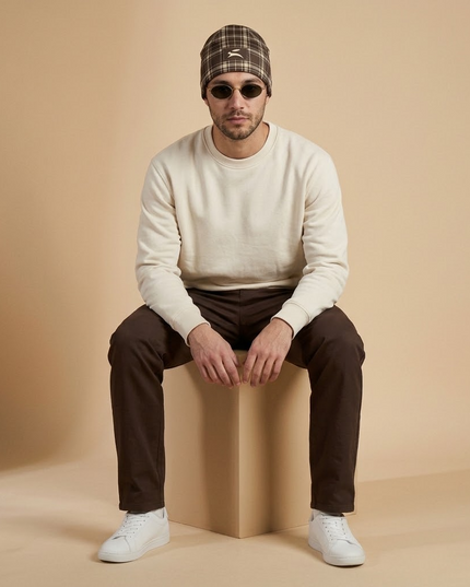 Heritage Tartan Beanie in Cocoa styled with a cream sweatshirt, seated full-length studio shot